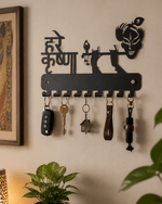 A black metal key holder wall mount featuring a silhouette of Lord Krishna and the Hindi phrase 'Hare Krishna', shown hanging on a beige wall with several keys and keychains attached. A framed picture, a lamp, and a green plant are visible in the room setting.