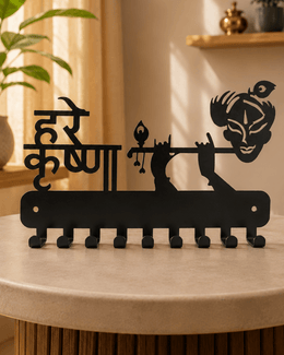 A black metal wall decor piece featuring the Hindi text 'Hare Krishna' and a stylized depiction of Lord Krishna, displayed on a round beige table in a warm, sunlit room.